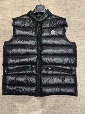 Moncler gui väst - Snygg svart dunväst från Moncler med glansig finish och klassisk logga på bröstet. Västen har ståkrage, två dragkedjeförsedda fickor och quiltad design. Perfekt för lager-på-lager och streetstyle.