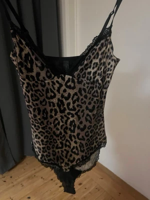 Leopardmönstrad spetsbody - Säljer en snygg leopardmönstrad body från Lindex med tunna axelband och svarta spetsdetaljer vid urringning och benslut. Materialet är mjukt och stretchigt, perfekt för dig som vill ha något både bekvämt och trendigt. Djurmönstret ger en cool vibe.