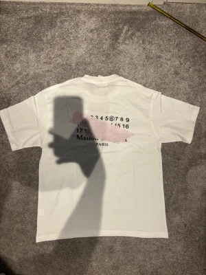 Vit Maison Margiela t-shirt - Vit t-shirt från Maison Margiela med tryck på både fram- och baksidan. Klassisk passform med korta ärmar och rund hals. Tillverkad i mjuk bomull och har diskreta svarta detaljer samt logotyptryck på bröstet.