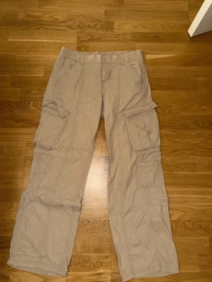 Beiga cargobyxor med fickor - Säljer ett par beiga cargobyxor med flera stora fickor på sidorna och avslappnad passform. Byxorna har resår i midjan och är tillverkade i ett lätt bomullstyg, perfekta för en chill och casual stil.