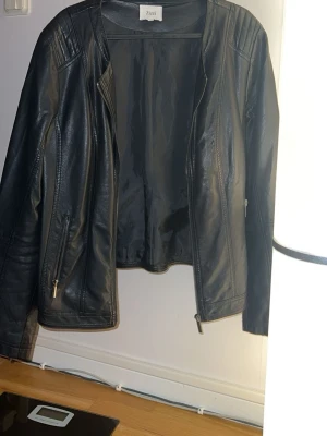 Svart skinnjacka från Zizzi - Snygg svart skinnjacka från Zizzi med dragkedjor på fickorna och detaljer vid axlarna. Jackan har en klassisk rak passform och stilrena sömmar längs ryggen och ärmarna. Perfekt för dig som vill ha en tidlös look.