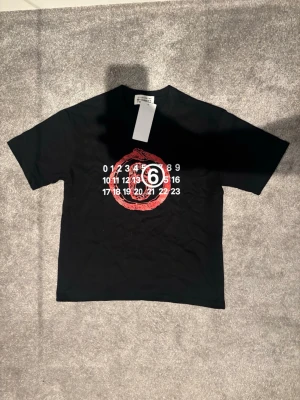 Maison Margiela t shirt - Cool svart t-shirt från Maison Margiela med tryck av siffror och en röd cirkel på bröstet. Klassisk passform med korta ärmar och rund hals. Perfekt för dig som gillar streetwear och unika designer.