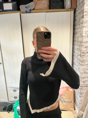 Svart stickad tröja med vit kant - Trendig svart stickad tröja med asymmetrisk design och vit kontrastkant längs ärm och nederkant. Tröjan har en liten polokrage och vida ärmar. Aldrig använt 
