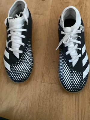 Två par fotbollsskor Adidas & Nike - Säljer två par fotbollsskor. Ett par är svartvita Adidas med klassiska ränder och rutmönster, och ett par är vita Nike med färgglada stänk och svarta detaljer. Båda paren har snörning och är tillverkade i syntetmaterial för bra grepp och känsla på planen.