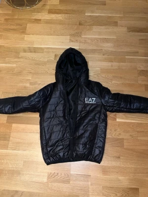 Svart pufferjacka med loggan EA7  - Säljer en svart pufferjacka med loggan EA7 med huva och dragkedja. Jackan har ett stilrent, quiltat mönster och logga på bröstet. Perfekt för kalla dagar och riktigt snygg till streetwear-looken.