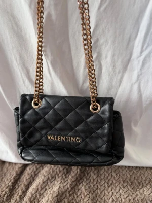 Svart quiltad handväska Valentino - Snygg svart handväska från Valentino med guld detaljer. Väskan har ett huvudfack med dragkedja och två innerfack för småsaker. Klassisk design i mjukt material. Väskan passar både till vardags och fest.