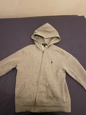 Grå hoodie från Polo Ralph Lauren - Säljer en stilren grå hoodie från Polo Ralph Lauren med dragkedja framtill och klassisk huva. Hoodien har två fickor och den ikoniska lilla loggan broderad på bröstet. Perfekt för en chill och avslappnad look.