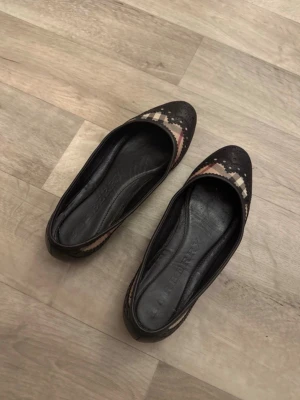 Svarta ballerinaskor med mönster - Säljer ett par svarta ballerinaskor från Burberry med klassiskt rutmönster i beige, rött och vitt framtill. Skorna har rund tå och dekorativa detaljer på ovandelen. Materialet är högkvalitativt läder, perfekt för dig som gillar stilrena och trendiga skor.