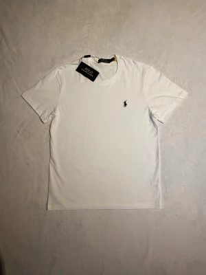 Vit t-shirt från Polo Ralph Lauren - Klassisk vit t-shirt från Polo Ralph Lauren med rund hals och korta ärmar. T-shirten har den ikoniska svarta polospelaren broderad på bröstet. Tillverkad i mjuk bomull för en skön känsla och clean look. Har även en annan exakt likadan!