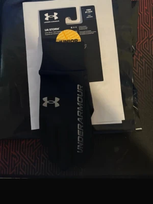 Svart under armor Storm handskar junior - Snygga svarta UA Storm handskar från Under Armour med logga och text i silver på ovansidan. Handskarna är tunna, smidiga och har greppvänlig insida. Perfekta för kyliga dagar och sportiga aktiviteter.
