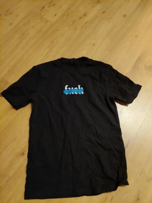 Svart t-shirt från FSBN med tryck - Svart t-shirt från FSBN med ett blåvitt tryck på bröstet. Klassisk passform och rund halsringning. Perfekt för dig som gillar enkel men snygg streetwear.