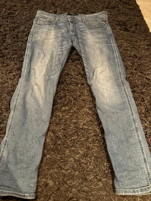Blå slim fit jeans från Replay 32/32 - Snygga blå slim fit jeans från Replay med klassisk femficksdesign och diskret slitning framtill. Jeansen har normal midja och är tillverkade i stretchigt denim för en bekväm passform. Perfekta för en trendig och avslappnad look.