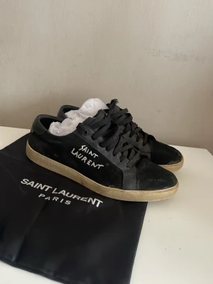 Svarta Saint Laurent sneakers - Svarta sneakers från Saint Laurent med vit logga på sidan och beige sula. Klassisk låg modell med snörning och ovandel i läder. Snygg och stilren design som passar till det mesta. Dustbag medföljer. Tar endast instabox.