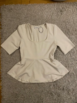 Vit peplum topp från Kappahl - Snygg vit topp från Kappahl med peplumskärning och fyrkantig ringning. Toppen har halvlånga ärmar och är tillverkad i ett mjukt material som ger en bekväm passform. Perfekt att styla med jeans eller kjol för en trendig look.