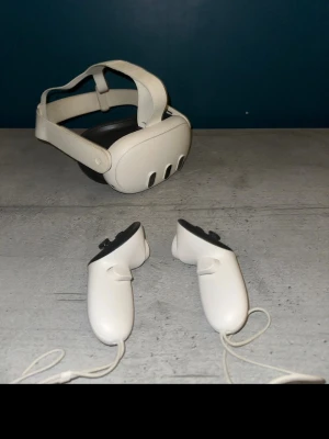 Meta Quest 3 VR-headset med kontroller och laddsladd - Säljer ett Oculus Quest 2 VR-headset i vitt med matchande handkontroller. Headsetet har justerbart band och modern design i plast. Perfekt för gaming och VR-upplevelser. Kontrollerna är ergonomiska och har handledsremmar för extra säkerhet.