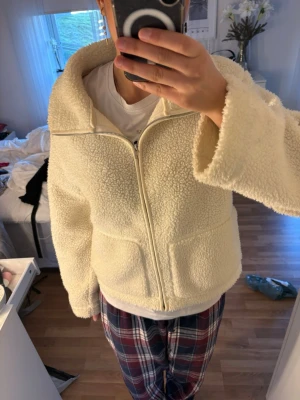 Krämvit teddyjacka med dragkedja - Mysig krämvit teddyjacka med stor krage och dragkedja framtill. Jackan har två stora fickor och är gjord i fluffigt, mjukt teddy-material som håller dig varm. Perfekt för kalla dagar och ger en avslappnad, trendig look.