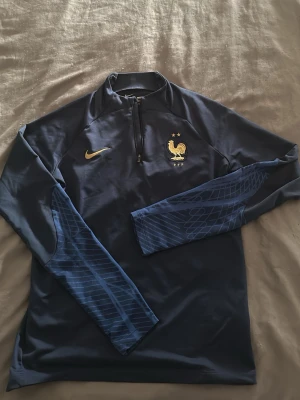 Blå Nike Frankrike träningströja - Mörkblå långärmad träningströja från Nike med Frankrikes landslagsmärke och guldigt Nike-logga på bröstet. Halvlång dragkedja framtill och mönstrade blå detaljer på ärmarna. Perfekt för fotbollsträning eller sportiga aktiviteter.