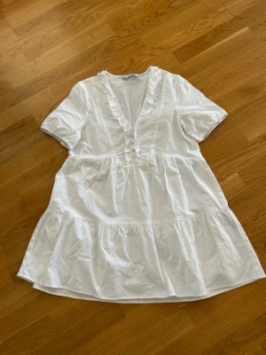  Ny Vit volangklänning från Zara - Superfin vit klänning från Zara med volangdetaljer längs v-ringningen och knappar framtill. Klänningen har korta ärmar och en luftig, kort modell med rynkade partier. Perfekt för varma dagar och enkel att styla med sneakers eller sandaler.