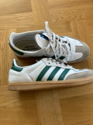 Adidas Samba vita och gröna sneakers - Klassiska Adidas Samba sneakers i vitt läder med gröna ränder och detaljer. Skorna har grå mocka framtill och gummisula. Ikonisk retrostil med låg profil och snörning. Perfekta för dig som gillar streetwear och sportig look.