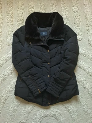 Svart pufferjacka från Stockh LM - Snygg svart pufferjacka från Stockh LM med fuskpälskrage. Lite sliten i ärmen, se sista bild. Inget som syns om man inte står o verkligen kollar