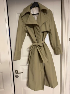 Beige trenchcoat från Tiger of Sweden - Snygg beige trenchcoat från Tiger of Sweden med knytskärp i midjan och stora knappar framtill. Jackan har klassisk krage och slät, glansig insida. 