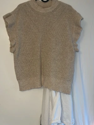 Beige stickad väst med volangärm från ONLY - Snygg beige stickad väst med volangdetaljer vid ärmarna och ribbad hög hals. Perfekt att styla över en skjorta eller t-shirt för en trendig lager-på-lager-look. Mjuk och skön kvalitet, loose fit och diskret mönsterstickning upptill.