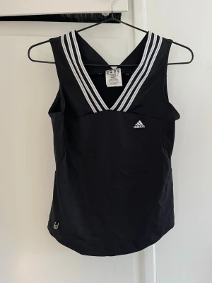 Svart sportlinne från Adidas - Snyggt svart sportlinne från Adidas med vita ränder längs V-ringad hals. Tillverkat i lätt och funktionellt material, perfekt för träning. Klassisk Adidas-logga framtill och ClimaLite-märke baktill. Ärmlös design för maximal rörelsefrihet.