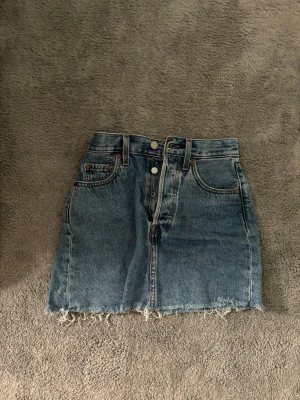 Blå jeanskjol från Levi's - Snygg blå jeanskjol från Levi's med klassiska fem fickor, knappar framtill och fransig kant nedtill. Perfekt för dig som gillar en avslappnad och trendig stil. Levi's-logga bak på linningen och slitna detaljer för extra edge.