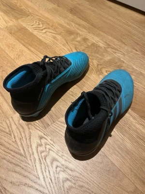 Adidas blå fotbollsskor med socka - Säljer ett par Adidas fotbollsskor i blått och svart med hög sockliknande krage och snörning. Skorna har mönstrad ovandel och dobbar för spel på gräs. Tillverkade i syntetmaterial och designade för bra grepp och kontroll på planen.