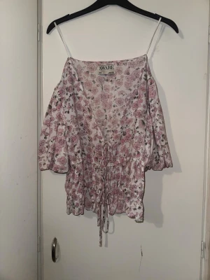 Blommig omlottopp från AWARE - Söt blommig omlottopp från AWARE (Vero Moda)med puffiga trekvartsärmar och snörning i midjan. Toppen har en v-ringad urringning och volangkant nedtill. 