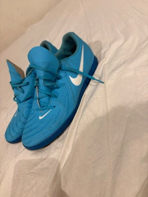 Blå Nike fotbollsskor inomhus - Säljer ett par blå Nike fotbollsskor för inomhusbruk med vit swoosh-logga på sidan. Skorna har snörning, låg profil och mönstrad ovandel i syntetmaterial för bra bollkontroll. Snygg sportig design som passar perfekt till futsal eller inomhusträning.