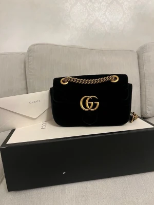 Svart sammetsväska från Gucci - Snygg svart handväska från Gucci i mjuk sammet med ikoniskt GG-spänne i guld framtill. Väskan har en kedjerem i guld med svarta läderdetaljer och en stilren, klassisk form. Perfekt för dig som vill ha en lyxig accessoar med statement-känsla.