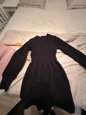 Svart stickad långärmad klänning - Mysig svart stickad klänning med lång ärm och rund hals. Klänningen har ett kabelstickat mönster upptill och är figurnära i midjan med en lätt utställd kjol. Perfekt för kyligare dagar och enkel att styla.