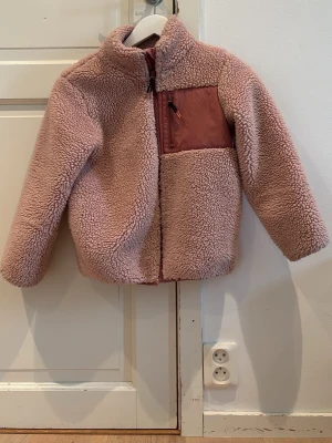 Rosa teddyjacka med bröstficka - Mysig rosa teddyjacka med hög krage och dragkedja framtill. Jackan har en kontrasterande bröstficka i nylon med dragkedja och detaljer i samma ton. Perfekt för kalla dagar och ger en trendig look med sin fluffiga struktur.