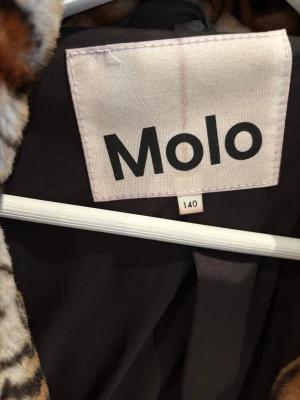 Leopardmönstrad pälsjacka från Molo - Snygg pälsjacka från Molo i storlek 140 med leopardmönster i bruna, svarta och beige toner. Jackan har mjukt fuskpälsmaterial och svart innerfoder. Perfekt för dig som vill sticka ut med en trendig look under vintern. Använda max 2-5 gånger!