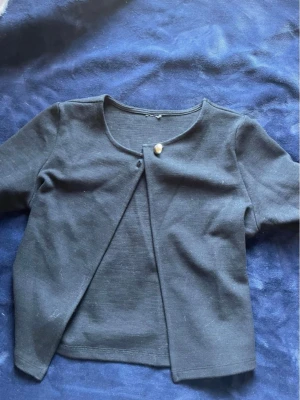 Svart kort kofta med knapp -  svart kort kofta med rund halsringning och en dekorativ guldfärgad knapp framtill. 