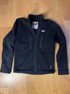 Fleec helly Hansen  - Svart fleecejacka från Helly Hansen med hög krage och dragkedja framtill. Två bröstfickor med dragkedja och diskret HH-logga på bröstet. Materialet är mjukt och värmande, perfekt för lager-på-lager under kyliga dagar.