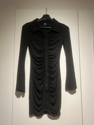 Svart klänning med knappar - Snygg svart klänning med långa ärmar och krage. Klänningen har knappar hela vägen framtill och ett rynkat parti som ger en cool effekt. Perfekt för dig som gillar en stilren men ändå unik look.