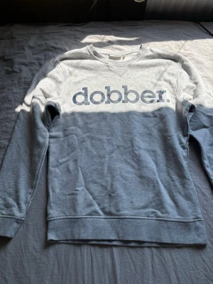 Grå sweatshirt från Dobber - Snygg grå sweatshirt från Dobber med stort tryck över bröstet. Tröjan har rund halsringning, ribbade muddar vid ärmslut och nederkant samt en avslappnad passform. Perfekt för en casual look och enkel att matcha med jeans eller joggers.