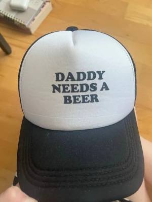 Svartvit truckerkeps med text - Säljer en svartvit truckerkeps med texten 'DADDY NEEDS A BEER' framtill. Kepsen har klassisk böjd skärm och mesh på sidorna för extra ventilation. Perfekt för dig som vill sticka ut med en rolig statement-accessoar.