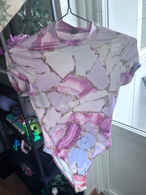 Rosa Guld Body - Säljer en kortärmad body med höghals i ett glansigt material. Bodyn har ett unikt mönster i lila, rosa och guldtoner som påminner om marmorerad sten. Perfekt för dig som vill sticka ut med färg och cool design.
