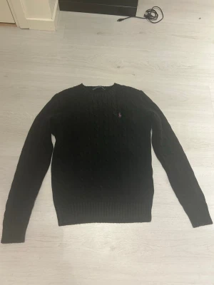 Svart kabelstickad tröja Polo Ralph Lauren - Svart kabelstickad tröja från Polo Ralph Lauren med klassisk rund halsringning och lila broderad logga på bröstet. Tröjan har långa ärmar och ribbade muddar. Perfekt för en stilren och tidlös look. Lite liten i storlek, står M men passar bättre som S.