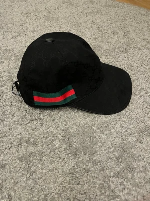 Svart keps med grön-röd rand - Snygg svart keps med diskret mönster och en klassisk grön och röd rand på sidan. Kepsen har böjd skärm och justerbar rem bak. Materialet ser ut att vara bomull eller canvas, perfekt för en trendig streetwear-look.
