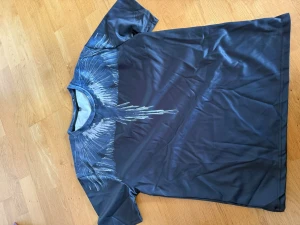 Svart t-shirt med vingeprint - Cool svart t-shirt med unikt vingeprint i vitt och grått över axlar och bröst. T-shirten har rund halsringning och korta ärmar. Materialet känns mjukt och lätt, perfekt för en avslappnad stil.