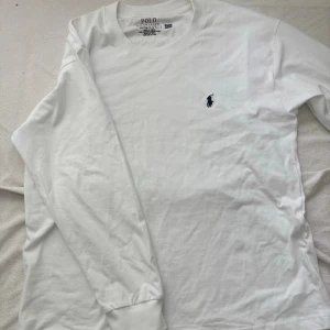 Vit långärmad t-shirt från Polo Ralph Lauren - Klassisk vit långärmad t-shirt från Polo Ralph Lauren med rund hals och den ikoniska broderade loggan på bröstet. Tillverkad i mjuk bomull för en skön känsla och clean look. Perfekt till jeans eller shorts för en enkel och stilren outfit.