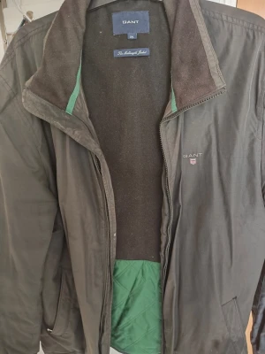 Svart vindjacka från GANT 2XL - Snygg svart vindjacka från GANT i modellen The Midlength Jacket. Jackan har dragkedja, ståkrage med brun insida och grönt innerfoder. GANT-logga broderad på bröstet och praktiska fickor med dragkedja. Perfekt för dig som vill ha en klassisk och stilren look.