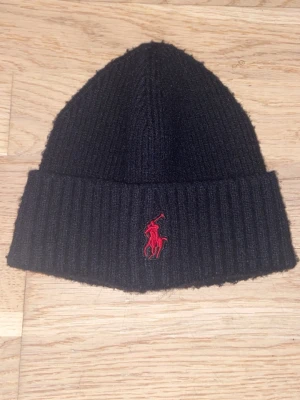 Ralph Lauren Mössa - Svart ribbstickad mössa från Polo Ralph Lauren med uppvikt kant och röd broderad logga framtill. Klassisk och enkel design som passar till det mesta. Mjuk och varm, perfekt för kyliga dagar.