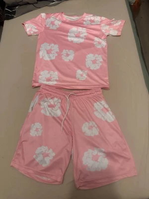 Denim tears set - Säljer ett tvådelat pyjamas-set i mjukt material. Setet består av en rosa t-shirt och matchande shorts med vita blommönster. Shortsen har resår och dragsko i midjan för extra komfort. Perfekt för mysiga hemmakvällar.