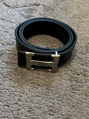 Svart bälte med silverfärgat H-spänne - Snyggt svart bälte med ett stort, silverfärgat H-format spänne och vita kontrastsömmar längs kanterna. Perfekt accessoar för att lyfta din outfit med en stilren och klassisk touch.