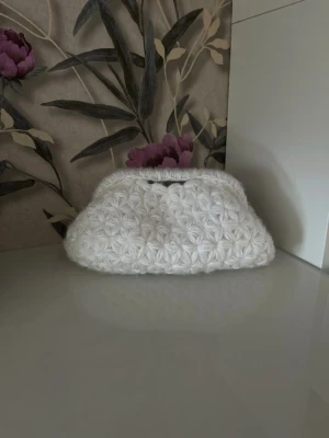 Vit virkad kuvertväska med blommönster - Säljer en superfin vit kuvertväska med virkat blommönster och mjuk struktur. Väskan har metallstängning upptill och ljusblått tygfoder på insidan. Perfekt för dig som vill ha en unik accessoar med vintagekänsla. Handvirkad av mig och är 18 cm bred💗